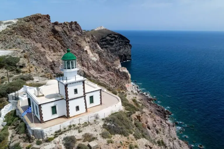 The lighthouse at Akrotiri seen from the air: a green-domed tower over a white keeper's house, right on the cliff edge above the sea.