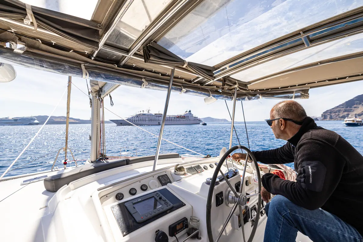 The skipper at the wheel under the bimini, a cruise ship at anchor across the water.
