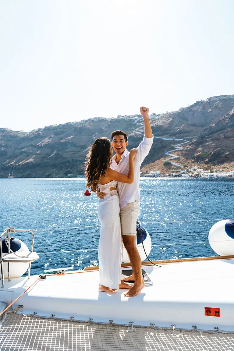 Two guests on the catamaran's foredeck, one with an arm raised, the caldera cliffs and a shoreline village behind them.