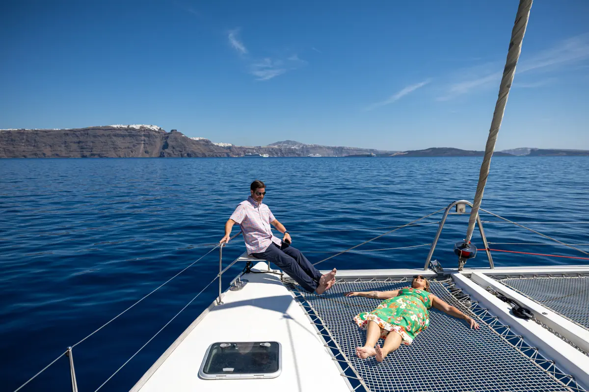 Two guests on the catamaran’s foredeck with the whole caldera rim running across the water behind them.