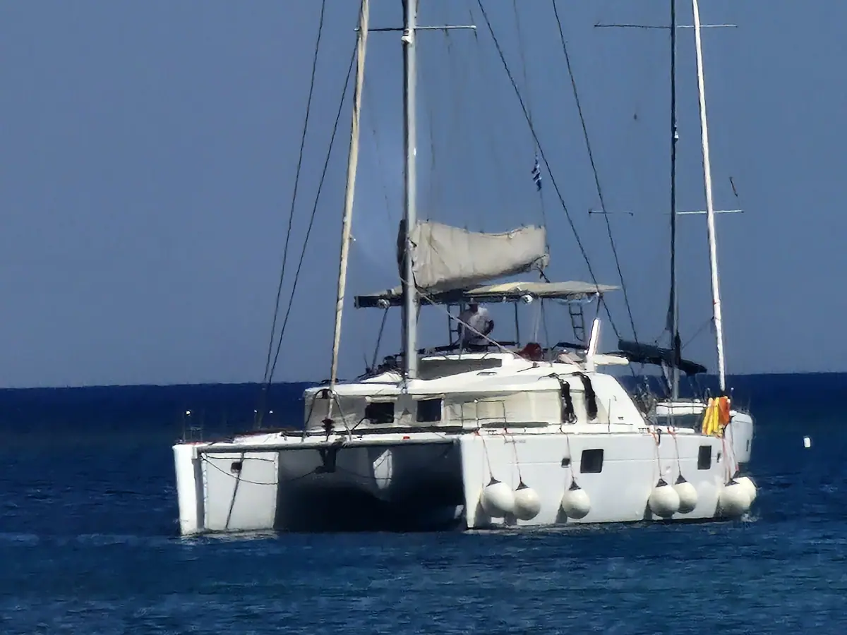 The catamaran seen beam-on at anchor, sails furled, on flat blue water.
