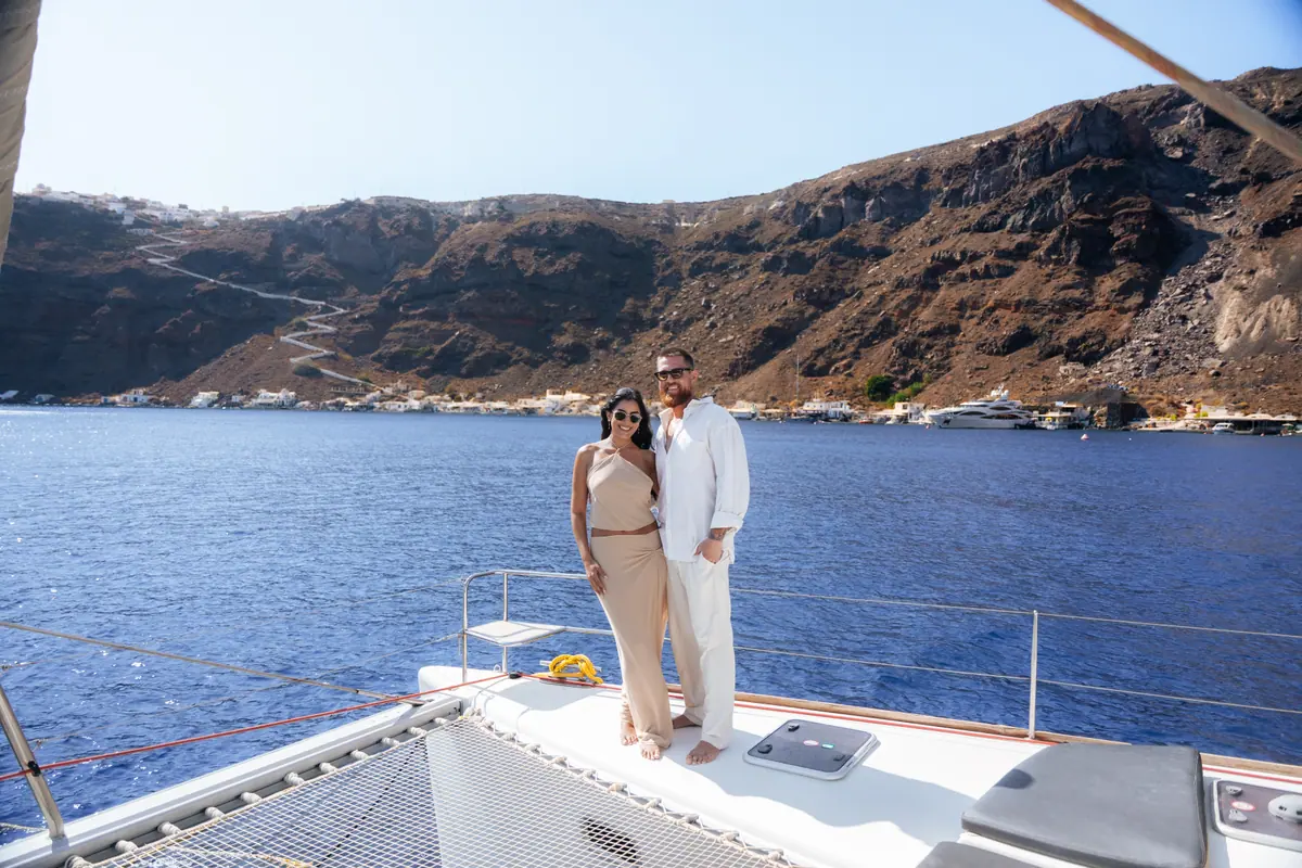 A couple standing at the bow, the red cliff of Ammoudi and the white houses of Oia above the water behind them.