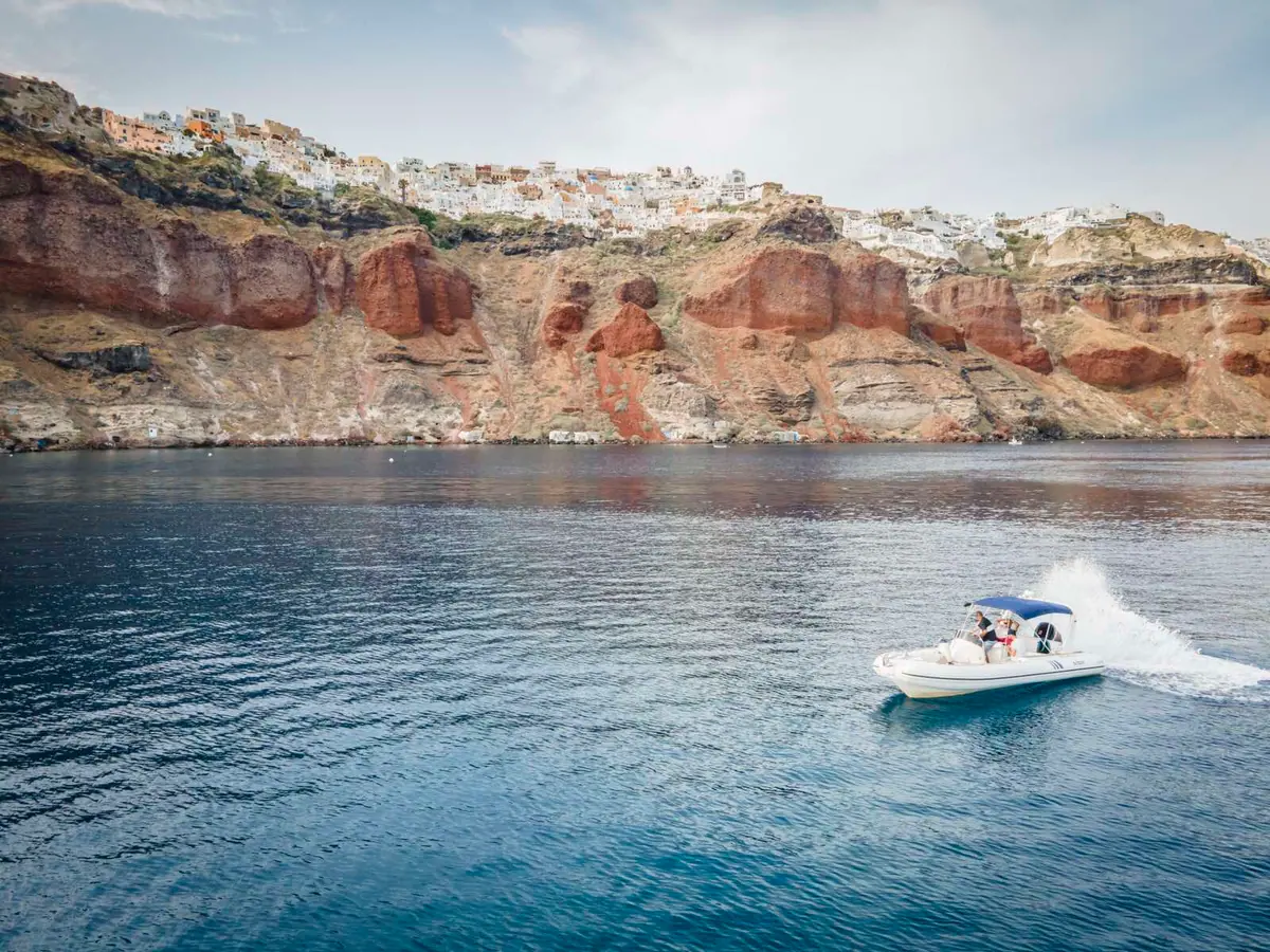The RIB running at speed with its wake behind it, below the red cliffs of the caldera.