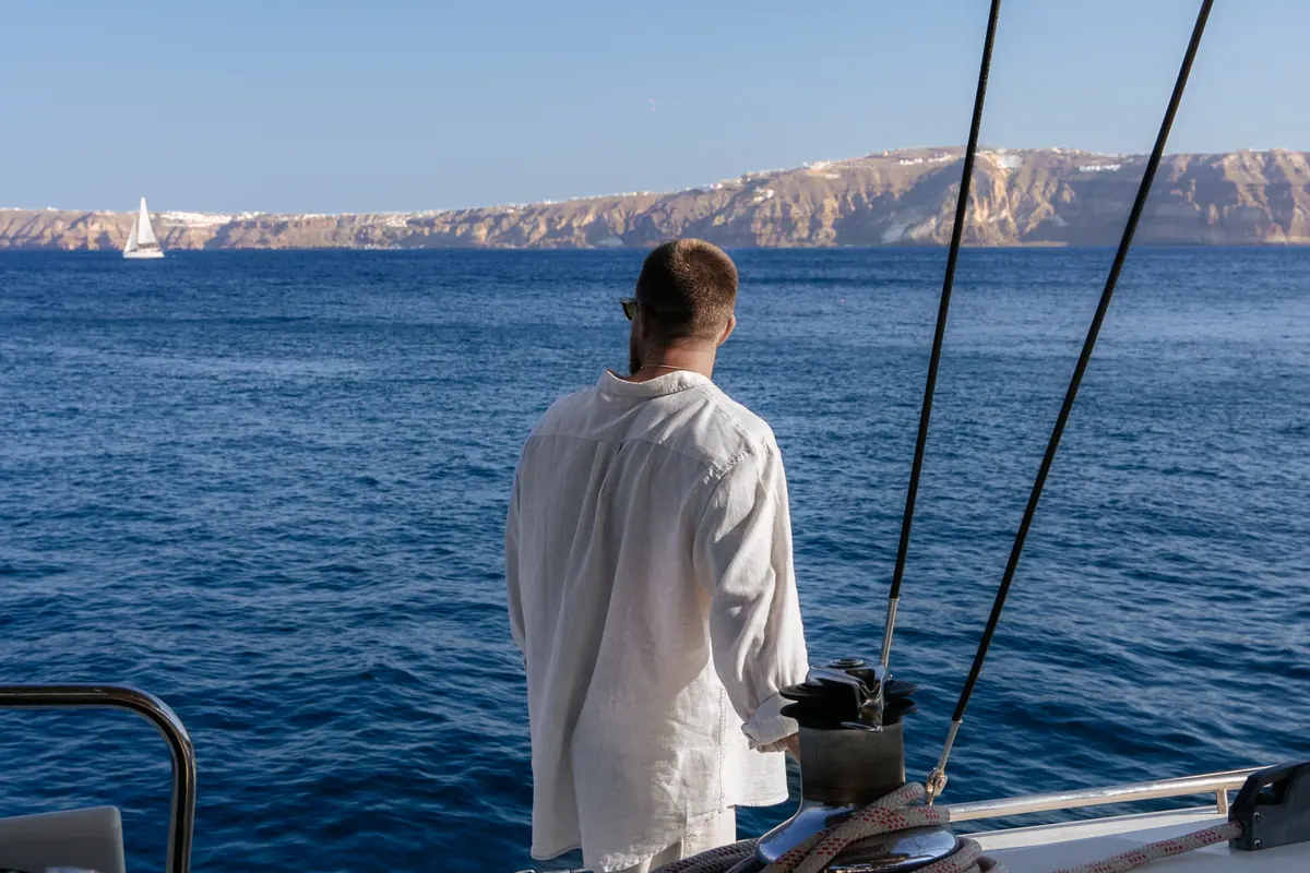 A guest at the rail under way, the caldera rim and a sail across the water.