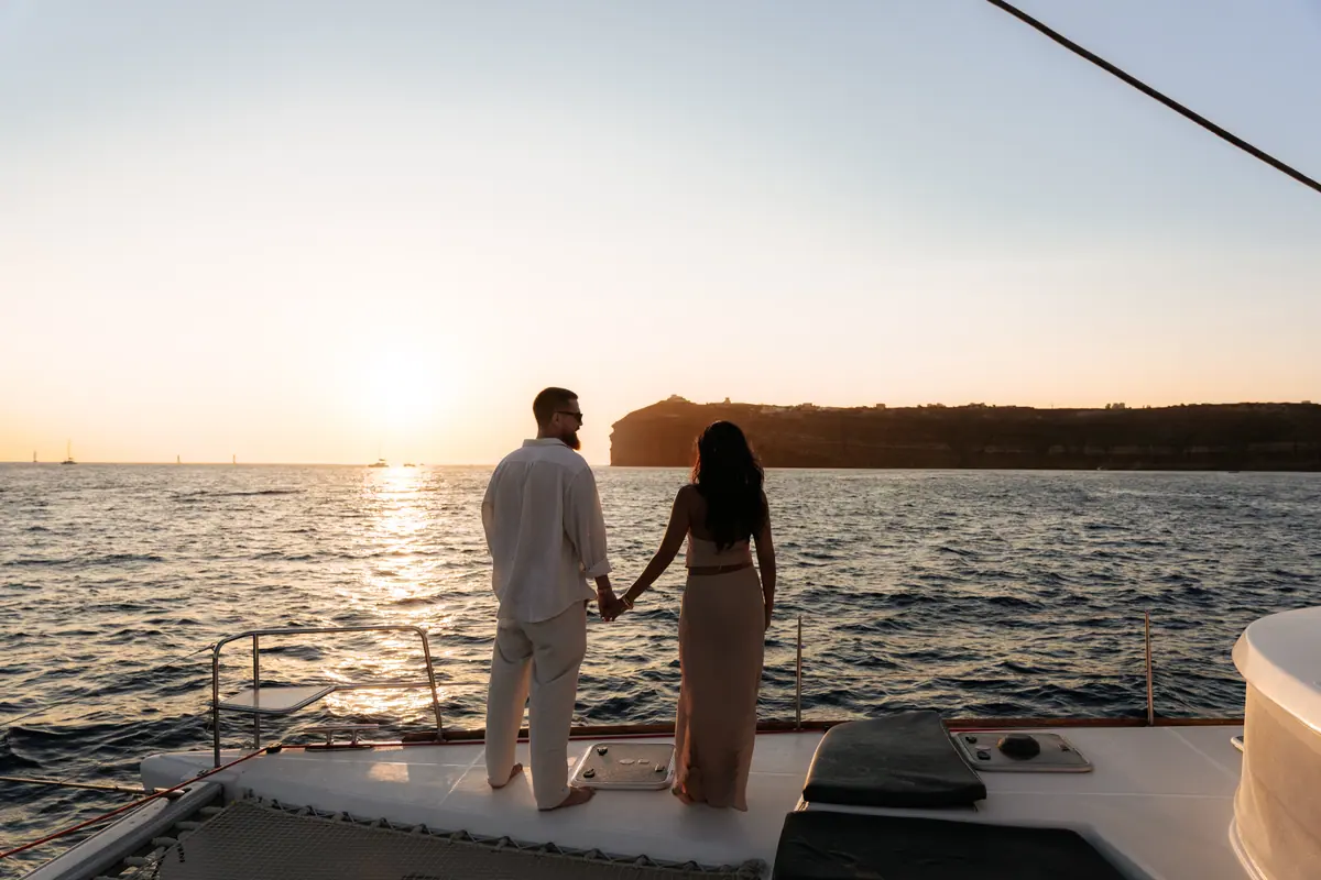 A couple hand in hand at the bow, the sun setting beside the Akrotiri headland.