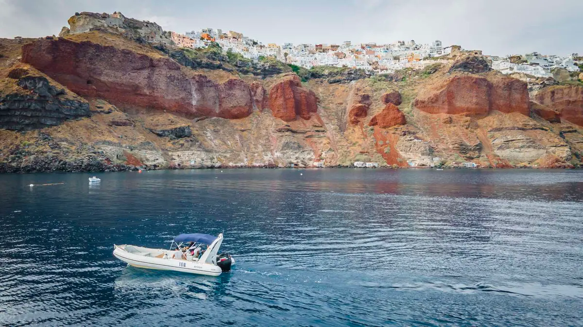 Le semi-rigide transporte des passagers sur une eau plate sous la falaise rouge d’Oia.