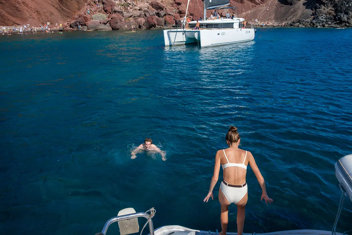 Red Beach from the water: the red cliff over the red shingle, a catamaran anchored off and swimmers in the bay.