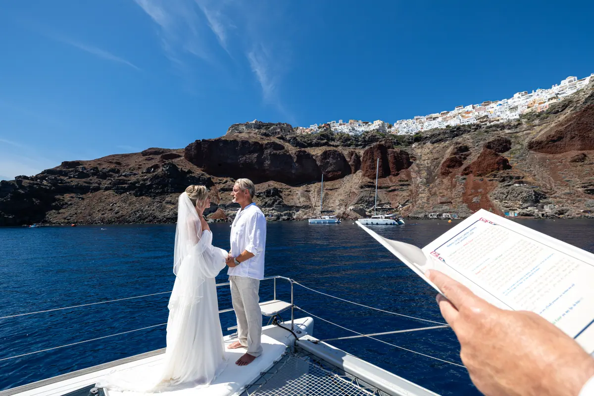 A wedding ceremony on the catamaran’s foredeck, the caldera cliff behind the couple.